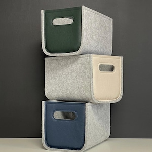 May include: Three stacked storage bins. The bins are gray felt with leather-like accents in green, cream, and navy blue. Each bin has a rectangular cutout for a handle. The bins are stacked against a dark gray background.