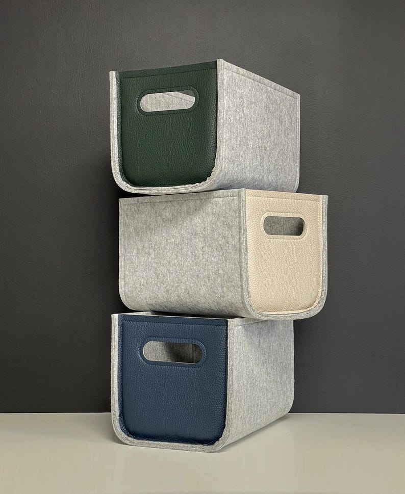 May include: Three stacked storage bins. The bins are gray felt with leather-like accents in green, cream, and navy blue. Each bin has a rectangular cutout for a handle. The bins are stacked against a dark gray background.
