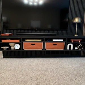 May include: A black entertainment center with multiple compartments and drawers. Two brown leather-like storage bins are visible. A gold-colored lamp and a clock are on display. The TV is mounted on a black wall.