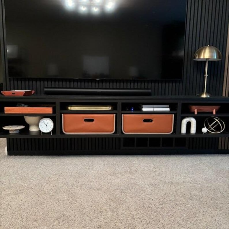 May include: A black entertainment center with multiple compartments and drawers. Two brown leather-like storage bins are visible. A gold-colored lamp and a clock are on display. The TV is mounted on a black wall.