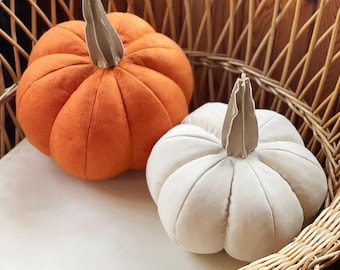 May include: Two plush pumpkins in a wicker chair. One is orange, the other is white. Both have tan stems. The pumpkins are sitting on a white cushion. The chair is made of woven wicker.