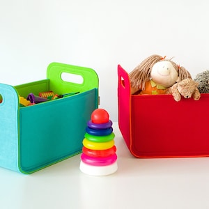 May include: Two colorful storage bins, one teal and the other red, sit side-by-side. The teal bin contains toys, while the red bin holds stuffed animals. A rainbow stacking toy is in front of the bins. The bins have handles.