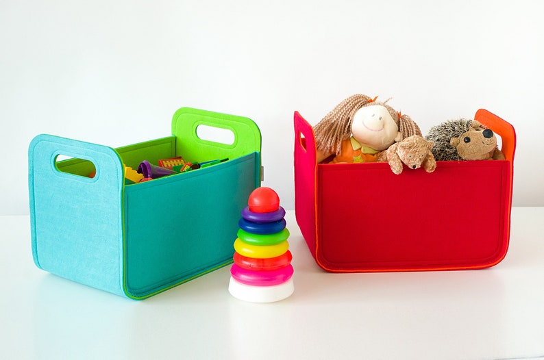 May include: Two colorful storage bins, one teal and the other red, sit side-by-side. The teal bin contains toys, while the red bin holds stuffed animals. A rainbow stacking toy is in front of the bins. The bins have handles.
