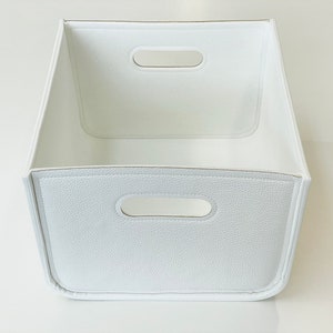 May include: A white storage bin with a textured exterior and two handle cutouts. The bin has a rectangular shape with a slightly trapezoidal design. The interior is smooth and white.