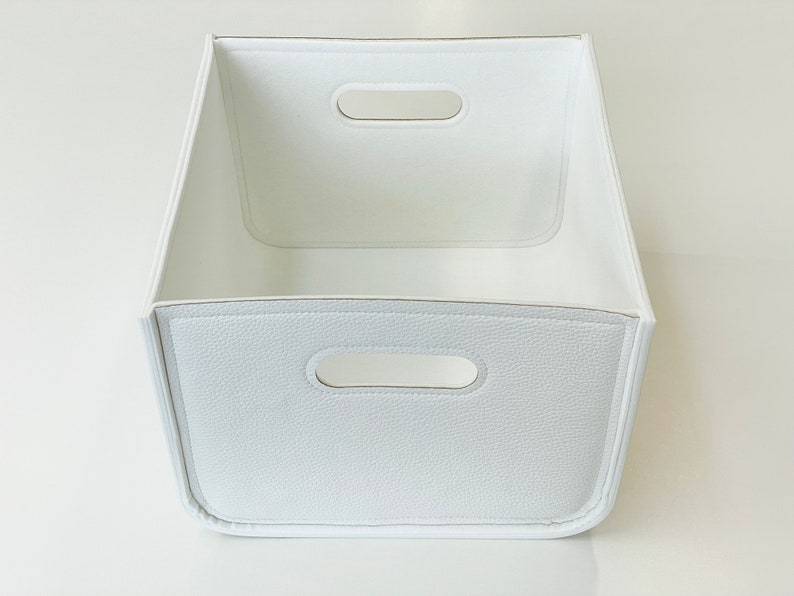 May include: A white storage bin with a textured exterior and two handle cutouts. The bin has a rectangular shape with a slightly trapezoidal design. The interior is smooth and white.