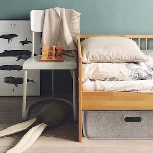 May include: A child's bedroom scene with a wooden bed, a white chair, and a whale poster. The bed has a light-colored duvet and pillows. A gray storage bin is under the bed. A stuffed animal with long ears lies on the floor.