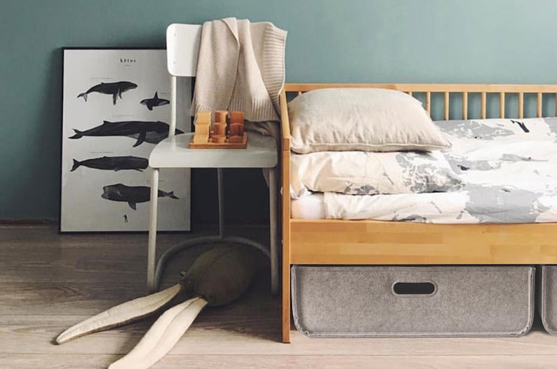 May include: A child's bedroom scene with a wooden bed, a white chair, and a whale poster. The bed has a light-colored duvet and pillows. A gray storage bin is under the bed. A stuffed animal with long ears lies on the floor.