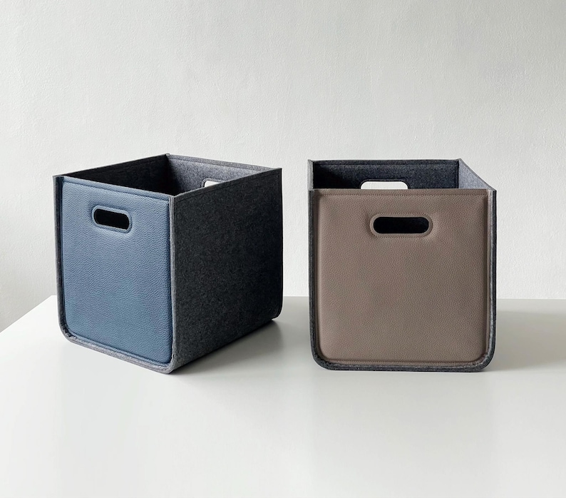 May include: Two square storage bins. One is blue with a gray felt interior and trim, and the other is taupe with a gray felt interior and trim. Each bin has a handle cutout on the front.