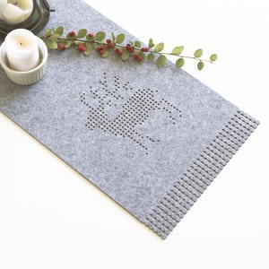 May include: Grey felt table runner with a dotted deer design and a fringed edge. The runner is decorated with a sprig of greenery and two candles.