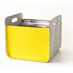 Op de afbeelding: Een grijze en gele opbergdoos van vilt met uitgesneden handvatten. De doos is gevuld met tijdschriften.