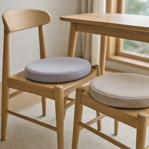 May include: Two wooden chairs with light brown cushions. The chairs have a simple design with straight legs and a rectangular seat. The cushions are round and have a soft, plush texture.