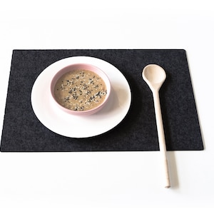 May include: A pink bowl with a beige soup topped with black and white sesame seeds sits on a white plate. A wooden spoon is on a dark gray placemat.