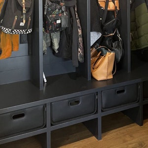May include: A black entryway bench with three storage drawers and three vertical compartments for hanging coats, bags, and backpacks. The bench is made of dark wood and has a modern design. Various bags and jackets are hanging in the compartments.