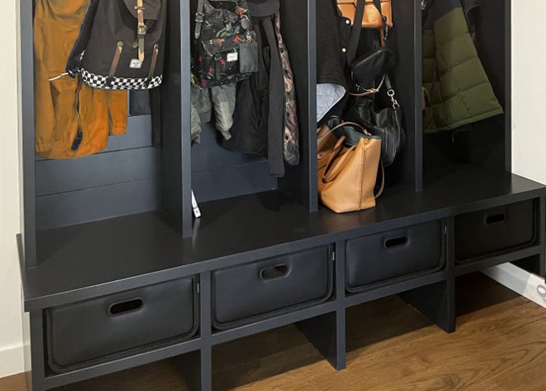 May include: A black entryway bench with three storage drawers and three vertical compartments for hanging coats, bags, and backpacks. The bench is made of dark wood and has a modern design. Various bags and jackets are hanging in the compartments.