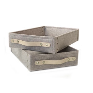 May include: Two grey felt storage boxes with leather handles and brass accents. The boxes are stacked on top of each other.