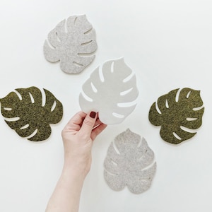 May include: Five felt leaf-shaped coasters, two are green and three are white. The coasters are arranged in a semi-circle on a white surface. A hand is holding one of the white coasters.