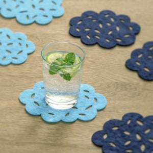 Puede incluir: Un vaso de agua con lima y menta descansa sobre un posavasos azul claro en forma de nube. Varios otros posavasos en forma de nube en azul claro y oscuro están sobre una superficie de madera. Los posavasos tienen diseños decorativos recortados.