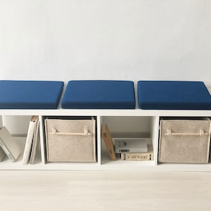 May include: A white bookshelf with three blue cushions on top. The bookshelf has three shelves with books and fabric storage bins.