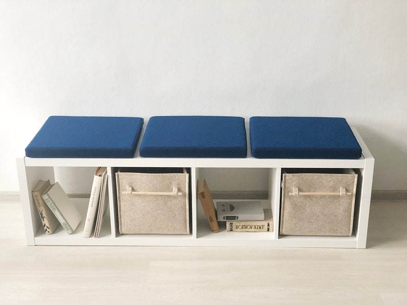 May include: A white bookshelf with three blue cushions on top. The bookshelf has three shelves with books and fabric storage bins.