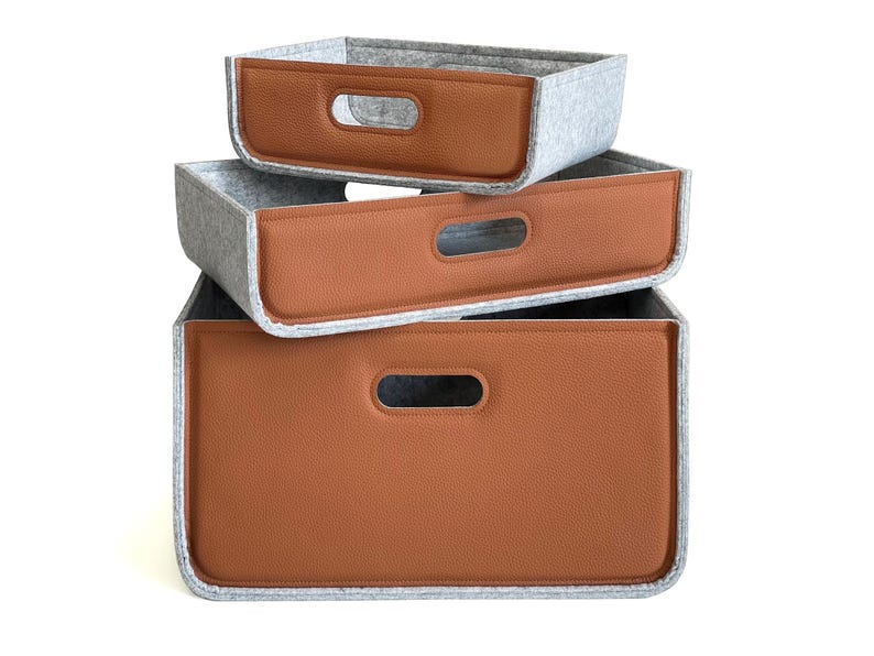 May include: Three rectangular storage bins stacked, each with a gray felt body and a brown faux leather front panel. The bins have a cutout handle and are designed for organizing items.