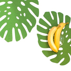 Op de afbeelding: Twee gele bananen in een groene monstera-bladschaal van vilt. De schaal is gevormd als een monstera-blad en heeft een witte achtergrond.