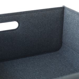 May include: A dark gray felt storage bin with a rectangular shape and a cutout handle. The bin has a simple, modern design and is made of a soft, textured material. The edges are neatly stitched.