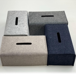 May include: A collection of four rectangular tissue box covers in various shades of gray and navy blue. Each cover has a rectangular opening on the top for tissue dispensing. The covers are made of a felt-like material.