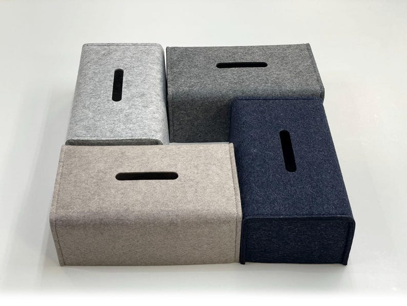 May include: A collection of four rectangular tissue box covers in various shades of gray and navy blue. Each cover has a rectangular opening on the top for tissue dispensing. The covers are made of a felt-like material.