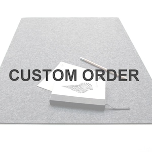 Op de afbeelding: Een grijze vilten mat met de woorden "CUSTOM ORDER" in het zwart. Een klein notitieboekje met een schelpenillustratie, een potlood en een stuk papier liggen op de mat.