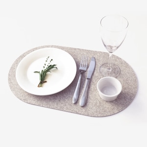 May include: A light gray oval felt placemat with a white plate, a small white bowl, a fork, a knife, and a clear wine glass. A sprig of green herbs sits on the plate.