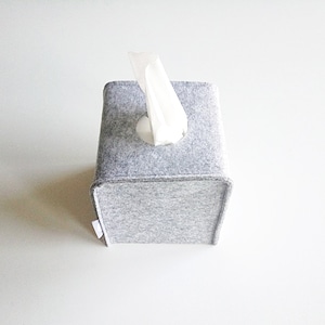 May include: A square, light gray felt tissue box cover with a white tissue protruding from the top. The box has a simple, modern design and is made of a soft, textured material. The tissue box is a neutral color and would fit in any room.