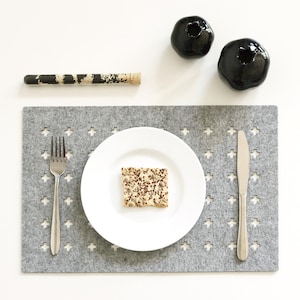 May include: A gray felt placemat with a white plate, a silver fork, and a silver knife. The plate has a small, square piece of bread on it. There are two black ceramic vases in the background.