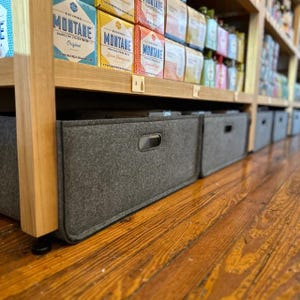 May include: A row of gray fabric storage bins with handle cutouts sit on a wooden floor beneath shelves stocked with beverage boxes. The bins are rectangular and have a simple, modern design.