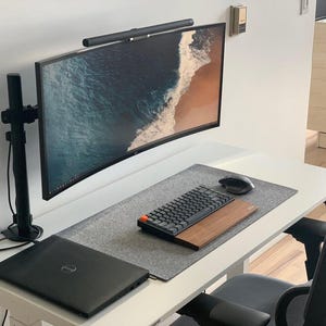 以下が含まれることがあります： 白い机に、湾曲したモニター、黒いキーボード、マウス、ラップトップが置かれています。モニターはアームに取り付けられており、上部にライトバーが付いています。机には、灰色のマウスパッドと木のキーボードトレイも付いています。