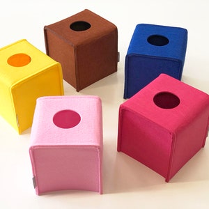 May include: A collection of six square tissue box covers in various colors: yellow, brown, blue, pink, and fuchsia. Each box has a circular opening on top. The covers are made of a soft, felt-like material.