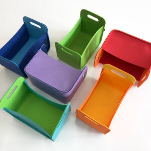 May include: Six colorful felt storage bins in blue, green, red, orange, purple, and lime green. Each bin has a rectangular shape with a handle on each side. The bins are arranged on a white surface.