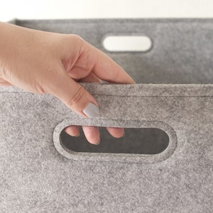 May include: Close-up of a gray felt storage bin with a rectangular handle cut-out. The bin is made of a soft, textured material. The handle is oval-shaped, and the edges are neatly finished. The overall design is simple and functional.