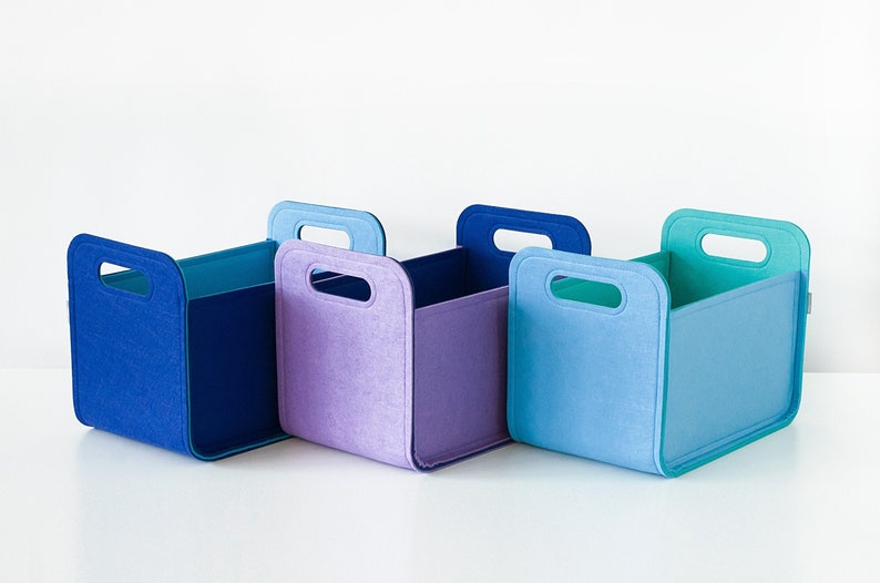 May include: Four colorful felt storage bins in shades of blue and purple. Each bin has a rectangular shape with a handle cut out at the top. The bins are designed for organizing and storage.