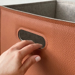 May include: A close-up of a storage bin with a brown, textured exterior and a gray interior. The bin has a rectangular shape with cut-out handles. The material appears to be faux leather. The bin is empty.
