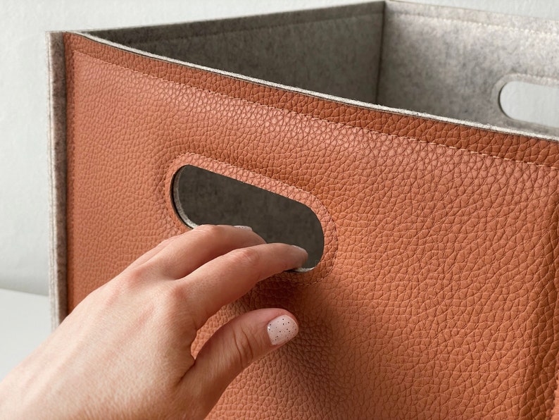 May include: A close-up of a storage bin with a brown, textured exterior and a gray interior. The bin has a rectangular shape with cut-out handles. The material appears to be faux leather. The bin is empty.