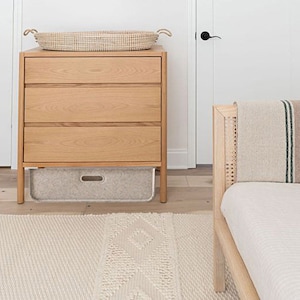 May include: A light wood dresser with three drawers and a woven basket on top. A gray storage bin sits below the drawers. A woven rug and a bed frame with a striped blanket are also visible.