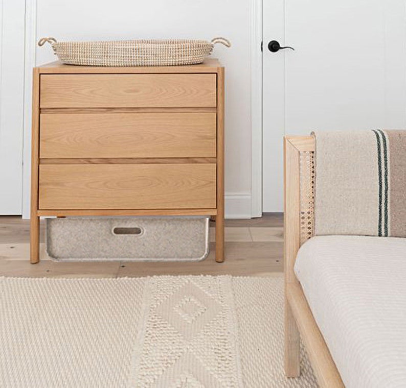 May include: A light wood dresser with three drawers and a woven basket on top. A gray storage bin sits below the drawers. A woven rug and a bed frame with a striped blanket are also visible.