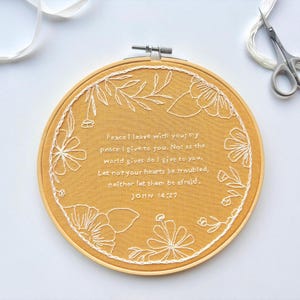 May include: A circular wooden embroidery hoop containing mustard yellow fabric with white thread embroidery. The design features a floral border surrounding the text: Peace I leave with you; my peace I give to you. John 14:27.