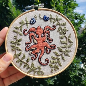 May include: A circular wooden embroidery hoop containing a piece of light-colored fabric. The fabric features a hand-stitched orange octopus centered within a circular wreath of green leaves and two small blue spiral shells at the top.