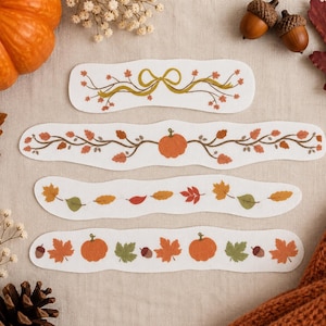 May include: Four white paper strips with wavy edges featuring autumn-themed illustrations. The designs include a yellow ribbon with leaves, a central orange pumpkin with vines, a row of colorful fallen leaves, and a pattern of pumpkins and acorns.