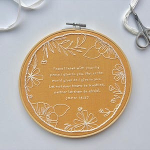 May include: A circular wooden embroidery hoop containing mustard yellow fabric with white thread embroidery. The design features a floral wreath surrounding the text: Peace I leave with you; my peace I give to you. Not as the world gives do I give to you. Let not your hearts be troubled, neither let them be afraid. JOHN 14:27.
