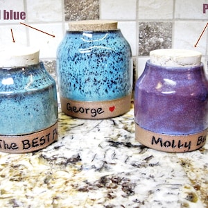 May include: Three ceramic jars with cork lids. The jars are teal blue, purple, and a light blue with brown speckles. The jars have personalized inscriptions on the bottom: "The Best", "Molly Be", and "George".