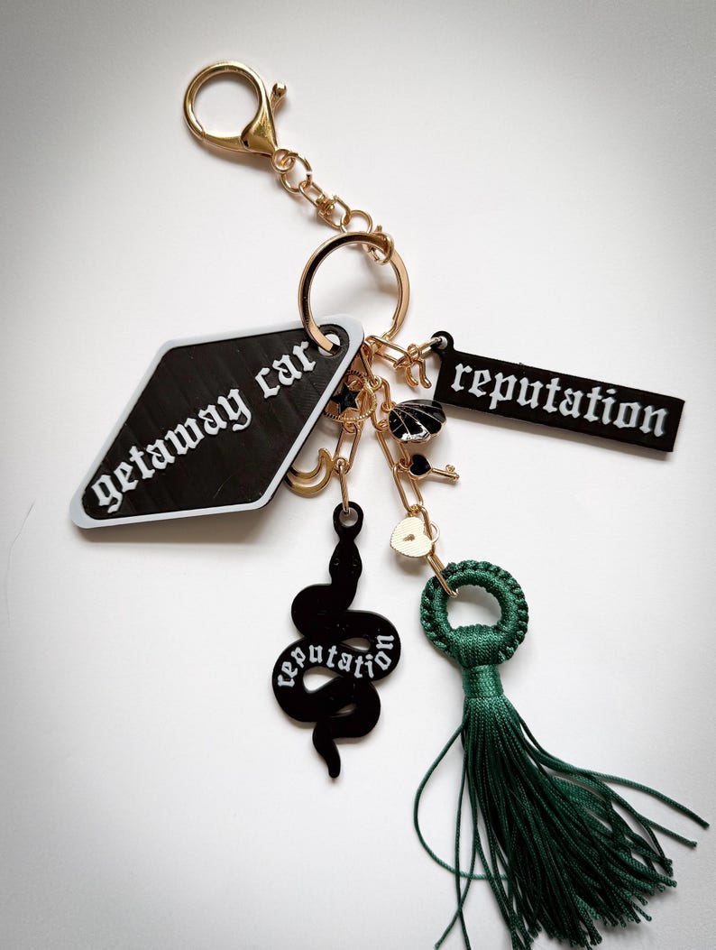 May include: A gold-tone keychain with multiple charms. Charms include a black diamond-shaped tag with "getaway car" text, a black rectangular tag with "reputation" text, a black snake, a key, and a green tassel. The keychain is on a white background.