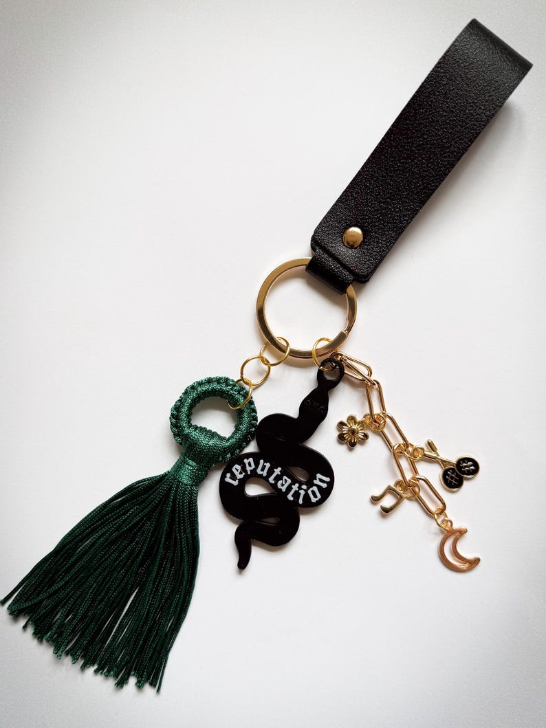 May include: A black leather keychain with gold-tone hardware and a dark green tassel. The keychain features a black snake charm with the word "reputation" and other gold-tone charms.