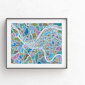 May include: A colorful watercolor map of a city with a river winding through it. The map is framed in a black frame and is hanging on a white wall.